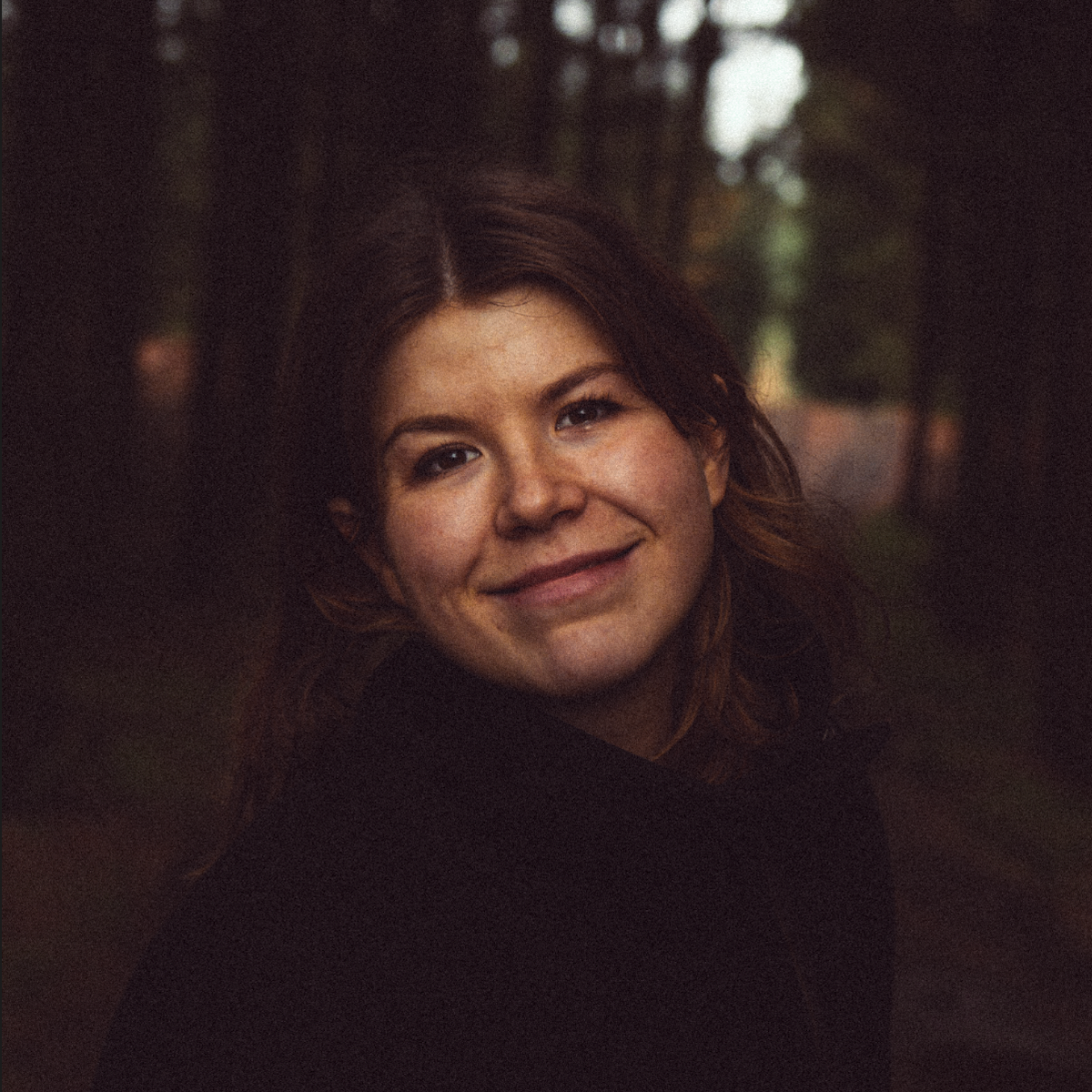 smilende mørkhåret kvinde i en mørk jakke i en mørk skov